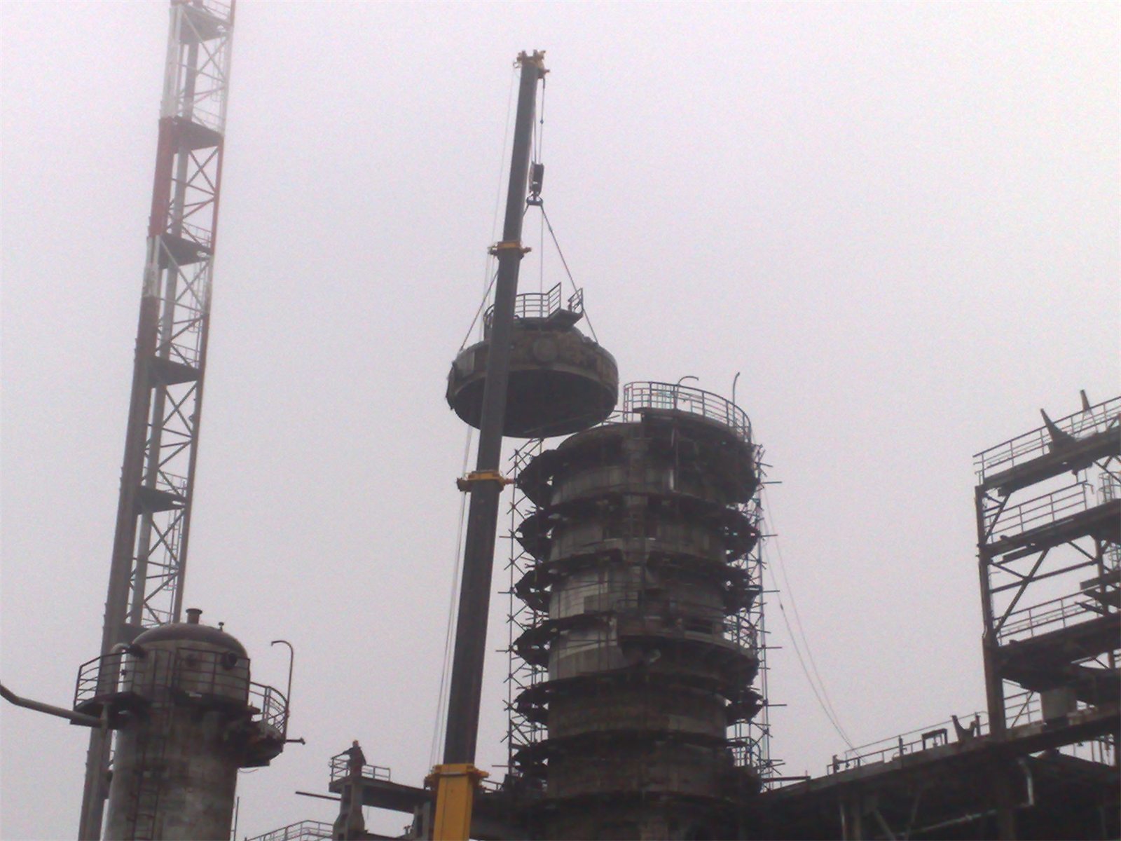 Section of a distillation column lifted by a mobile crane beside a column wrapped in scaffolding